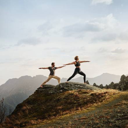 Yoga-Paar in den Alpen, Krieger-Pose auf Berggipfel mit Weitblick – Bewegung, Balance und Naturerlebnis.