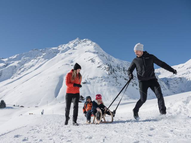 Kinder sitzten auf dem Schlitten und werden von Papa über die Piste gezogen und Mama läuft neben her