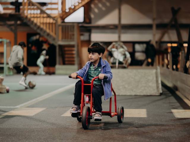 Ein Kind auf einem Dreirad im Indoospielplatz und andere Kinder sind im Hintergrund am spielen