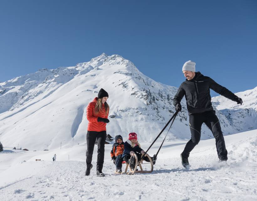 Kinder sitzten auf dem Schlitten und werden von Papa über die Piste gezogen und Mama läuft neben her