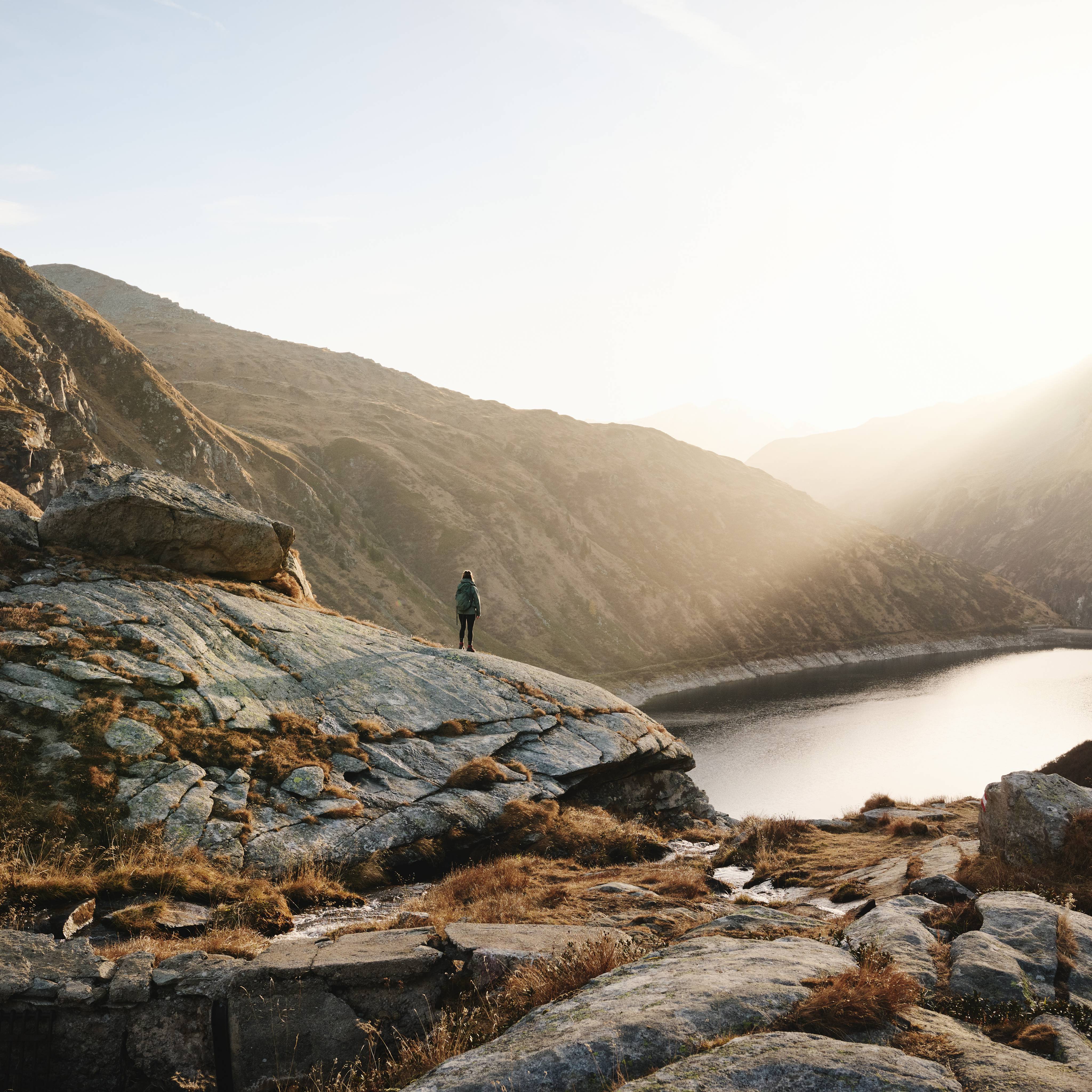 Eine Person steht auf Felsen, blickt auf den Bergsee während die Sonne über den Bergen aufgeht